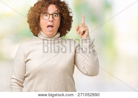 Beautiful middle ager senior woman wearing turtleneck sweater and glasses over isolated background pointing finger up with successful idea. Exited and happy. Number one.