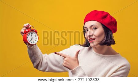 Young Woman In Red Beret With Alarm Clock. Woman Pointing On Clock Over Yellow Background. Girl Show