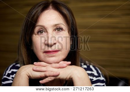 Close-up face of elderly woman sitting and propping chin with folded hands.