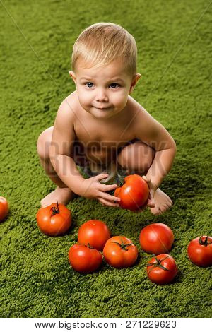 Close Up View Of Smiling Toddler Boy Sitting On Hunkers And Holding Tomato On Green Carpet