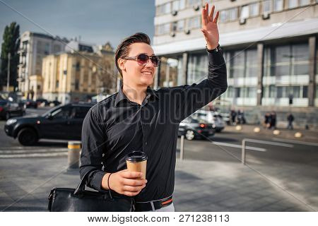 Chherful And Happy Young Man Walks On Street. He Waves With Hand. Guy Has Cup Of Drink And Bag.