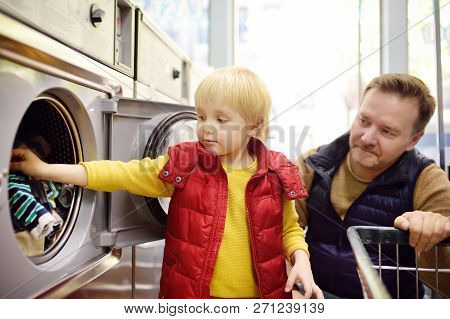 A Little Boy Loads Clothes Into The Washing Machine In Public Laundrette. Family In Public Laundry