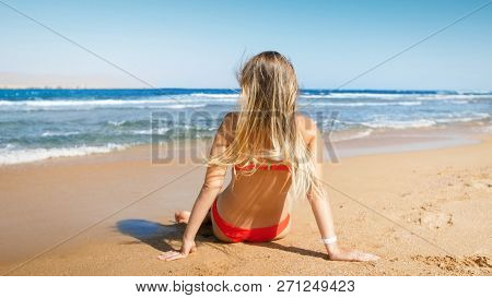 Rera View Image Of Young Woman In Red Bikini Sitting On The Beach And Looking At Sea