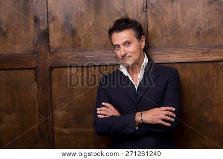 Calm Serious Middle Aged Man Standing Against The Wooden Wall And Thoughtfully Looking At You While 