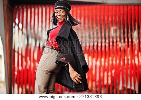 African American Fashion Girl In Coat And Newsboy Cap Posed At Street Against Red Background.