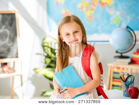 Little smiling blond girl standing near blackbord with red backpack and holding a notebook with pencils in the sunshine school classroom