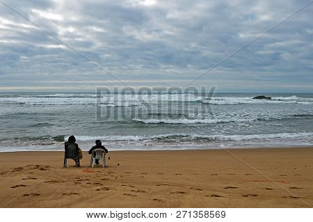 A Couple Siting On The Beach Praia De Odeceixe In South Portugal Near The Sea On The Cloudly Sky Bac