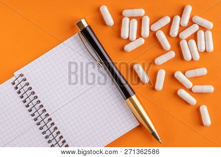 Medical Capsules And Pills Around A Blank Notebook With Pen On An Orange Background, Close-up, Top V