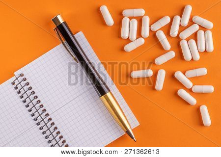 Medical Capsules And Pills Around A Blank Notebook With Pen On An Orange Background, Close-up, Top V