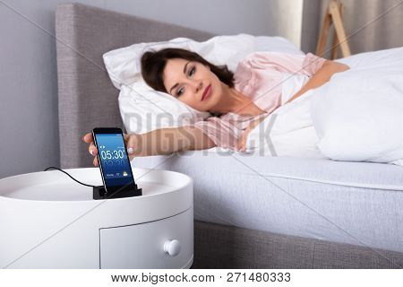 Beautiful Young Woman Lying On Bed Snoozing Alarm On Mobile Phone