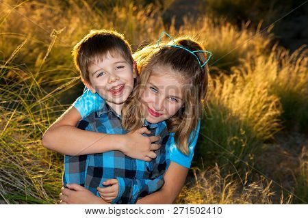A Young Girl Hugs Her Little Brother From Behind For A Portrait In Late Afternoon, Backlit Light.  T