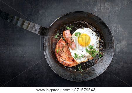Traditional fried Allgauer veal roast roll sausage slices with fried egg in a cast-iron skillet with spices as closeup on a black board 