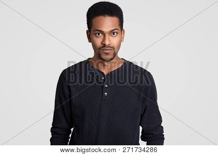 Indoor Shot Of Serious Self Confident Dark Skinned Young Man With Curly Short Hair, Looks With Stern