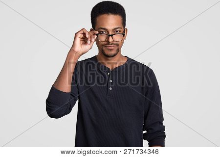 Waist Up Shot Of Handsome Self Confident Young Male Manager, Keeps Hand On Rim Of Spectacles, Wears 