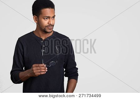 Studio Shot Of Black Male Director Focused Thoughtfully Aside, Holds Spectacles In Hands, Being Deep