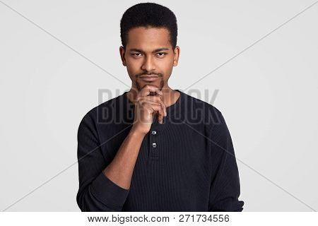 Headshot Of Self Assured Confident Serious Male Holds Chin With Hand, Has Beard, Dressed In Casual C