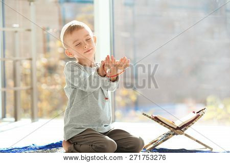 Little Muslim Boy With Misbaha And Koran Praying On Rug Indoors