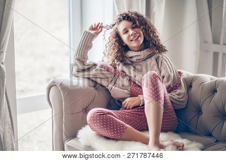Hipster teenage girl with curly hair wearing beige knitted sweater and pink polka dot jumpsuit relaxing on a couch indoor