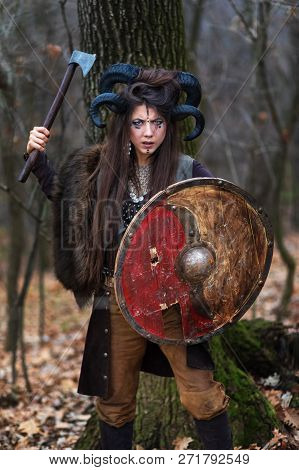 Outdoor Portrait Of Beautiful Viking Woman Warrior With Blue Eyes Wearing Ram Horns Holding Ax And S