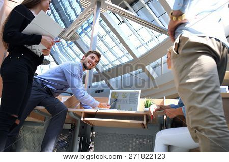 Group Of Young People In Casual Wear Sitting At The Office Desk And Discussing Something While Looki