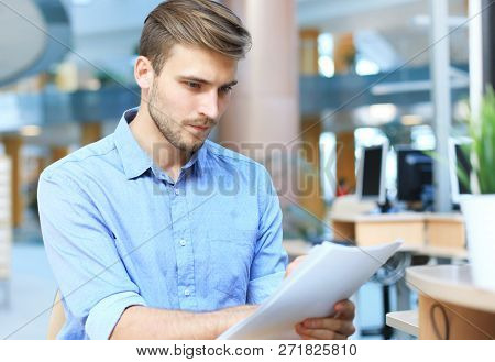 Young Businessman Reading Paperwork At Desk In Office.