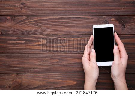 Female Hands Holding Mobile Phone With Blank Screen On Brown Wooden Table. Smartfone White