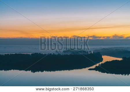 Early Blue Sky Reflected In River Water. Riverbank With Forest Under Predawn Sky. Yellow Stripe In P