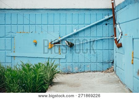 Blue Wall From Tiles With Cyan Closed Hatchway And Pipes Close Up. Background Of Wall Of Building Wi