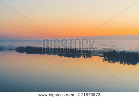 Broad Mystical River Flows Along Diagonal Shore With Silhouette Of Trees And Thick Fog. Gold Glow In
