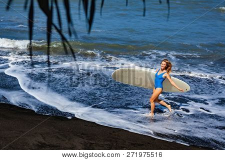 Sporty Girl In Bikini With Surf Board Walk On Black Sand Beach. Young Surfer Woman Run By Water Pool