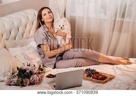 A Girl In Pajamas Sits In Bed At Night With Her White Dog Watching A Laptop And Eating Sweets.girl W