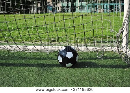 Black Soccer Ball In Goal Net On A Green Lawn On A Sunny Summer Day. The Goal Net Is Dirty And Torn.