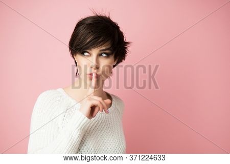 Beautiful young woman with long shot dark hair wears casual white sweater shows hush sign and looking up at left corner. Pretty lady asks not to tell her secret anybody.