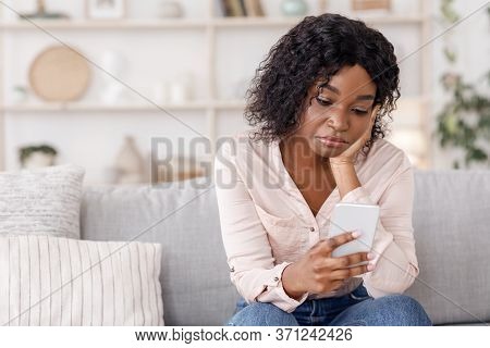 Waiting For His Call. Depressed Black Woman Looking At Cellphone With Sad Face, Sitting On Couch At 