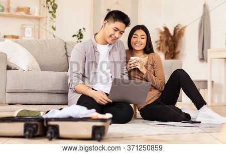 Isolation After Trip. Smiling Asian Couple Using Laptop Sitting On Floor Near Unpacked Suitcase At H