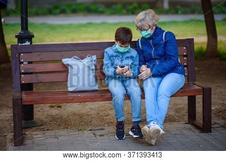 Grandmother With Grandson Outdoors In Park In Medical Masks Sit On A Bench With Smartphone. Boy Show