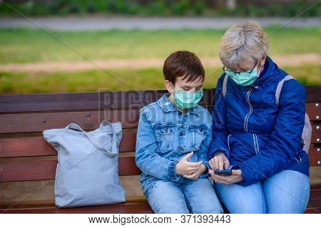 55 Years Old Pensioner Woman And A Boy In A Medical Masks Sit On A Park Bench With A Smartphone/ Wom