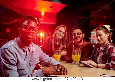 Happy young man of African ethnicity in casualwear looking at you while sitting by table with his intercultural friends having drinks in bar