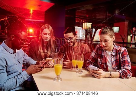 Group of four young intercultural friends in casualwear using mobile gadgets while sitting by table in sports bar and having drinks