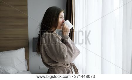Positive Girl With Loose Flowing Hair In Beige Bathrobe Drinks Tasty Coffee Standing Near Window In 