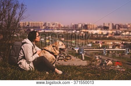 A Girl Sits Hugging A Labrador Retriever On A Mountain Against The Background Of The City. Vacation 