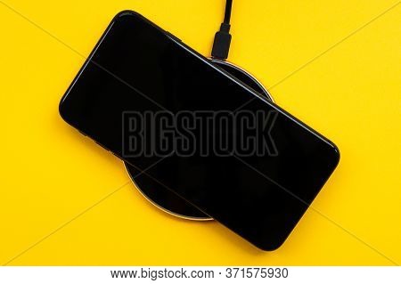 The Black Mobile Phone Is On Wireless Charging, On A Yellow Background. Modern Technologies, Electro