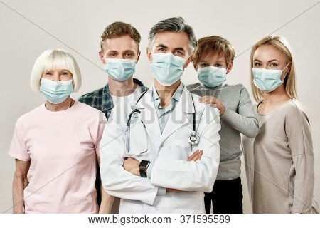 Family Physician. Portrait Of Mature Male Doctor And Family Of Four Wearing Medical Protective Masks