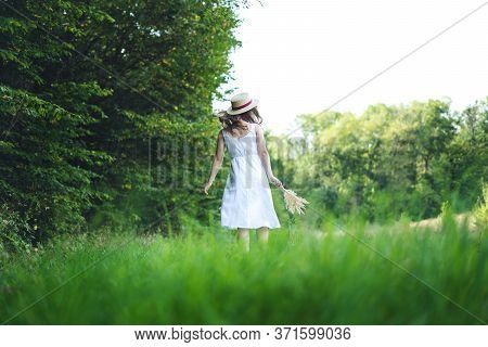 Young Woman Walking In A Green Field. Young Girl In A White Dress And Hat. Girl In A Dress And A Sum