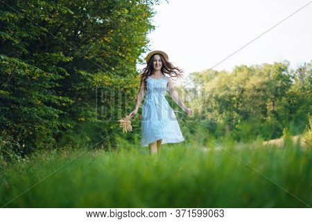 Young Woman Walking In A Green Field. Young Girl In A White Dress And Hat. Girl In A Dress And A Sum