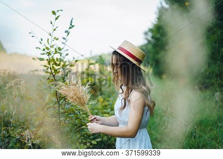 Smiling Girl In A Straw Hat Collects Flowers. Girl In A Summer Dress In The Tall Grass. Woman In A G