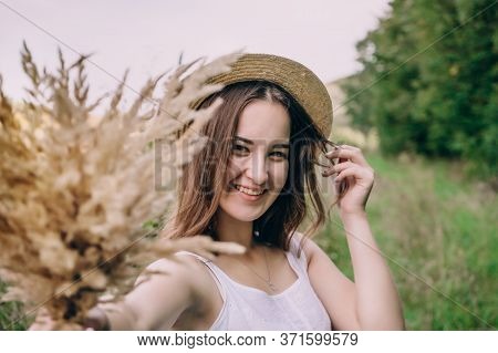 Happy Young Woman In A White Dress In A Straw Hat. Smiling Girl With A Bouquet Of Dried Flowers. Gir