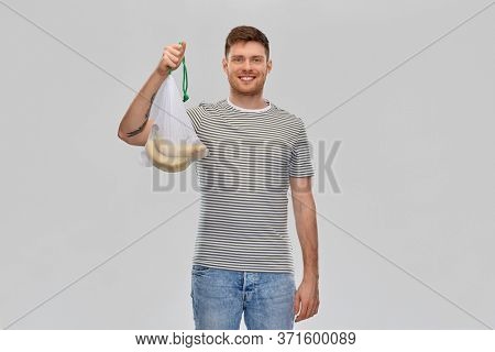 food, sustainability and eco living concept - smiling young man in striped t-shirt holding reusable string bag with bananas over grey background