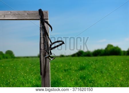 The Black Leather Snaffle Bridle Is Hanging On The Wooden Hitching Post On Against The Landscape.