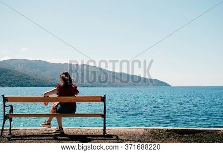 Unrecognisable Woman Seen From Behind Sitting On A Bench On The Seaside. Mountains And Endless Sea I